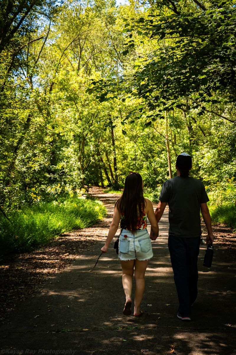 A couple walks hand in hand away down a dappled, tree-canopied path in summer light.