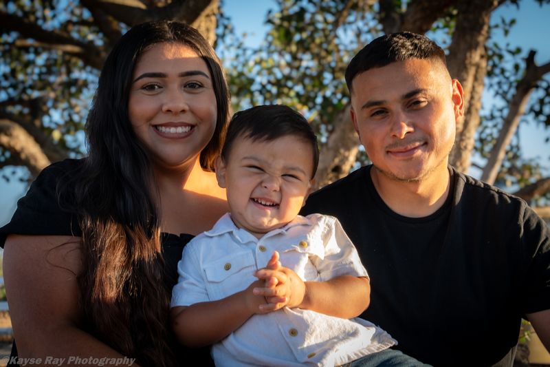 A toddler bursts into laughter between his smiling parents in warm golden-hour light under a tree.