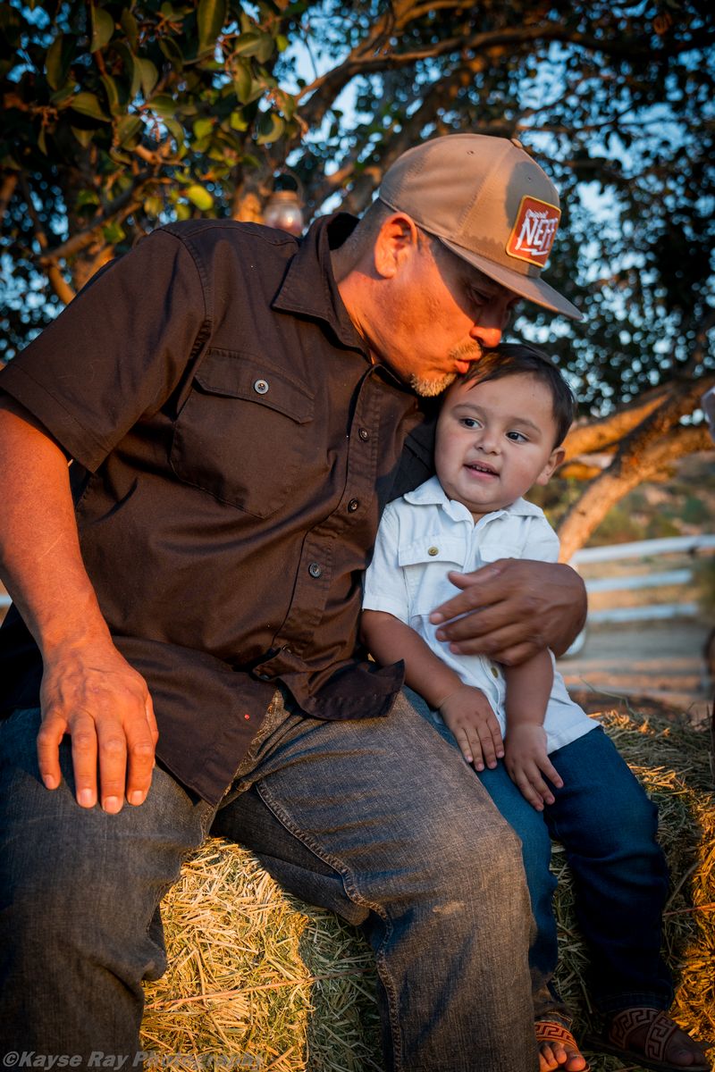 Father in a ball cap kissing the top of his young son's head as they sit together on a hay bale at sunset.