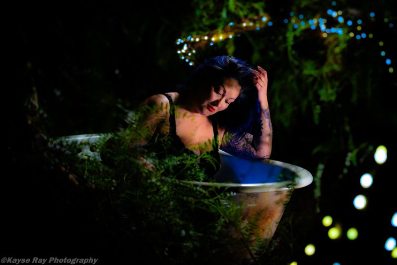 Woman running a hand through her dark hair in a moonlit tub, eyes closed against a bokeh of colored lights.