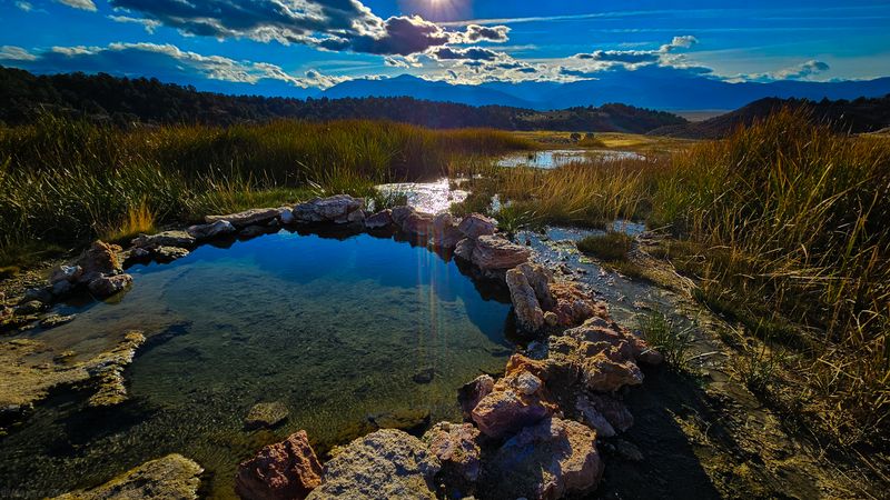 Rock-rimmed hot spring pool reflecting a bright sky above golden meadow grasses and distant mountains