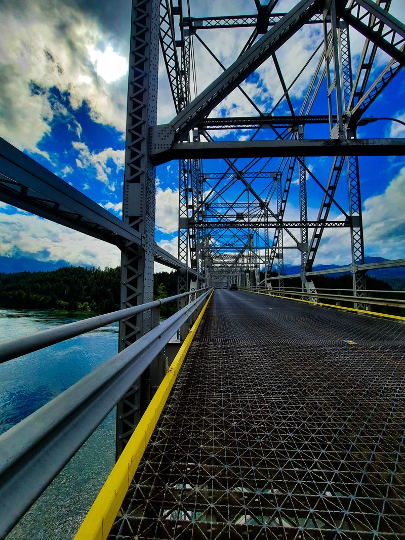 Steel truss bridge deck receding into the distance with yellow railings and a river far below